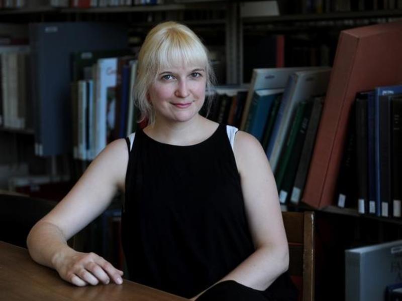[Image description: Donna Kozloskie in a black sleeveless shirt standing in front of bookshelves smiling at the camera. Her right arm is resting on the table in front of her and her left arm is placed in her lap.]