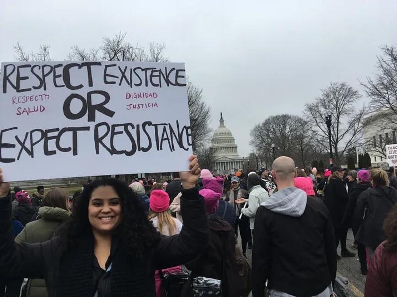 [image description: Emmeline Rodriguez in a marching crowd. She is facing the camera and smiling, holding a sign that says "Respect Existence OR Expect Resistance /  Respecto Salud OR Dignidad Jusitcia"]